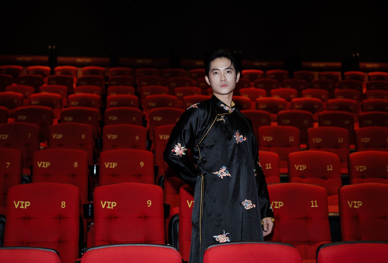 Portrait of an Asian man in traditional clothing standing in a VIP cinema with red seats and floral patterns.
