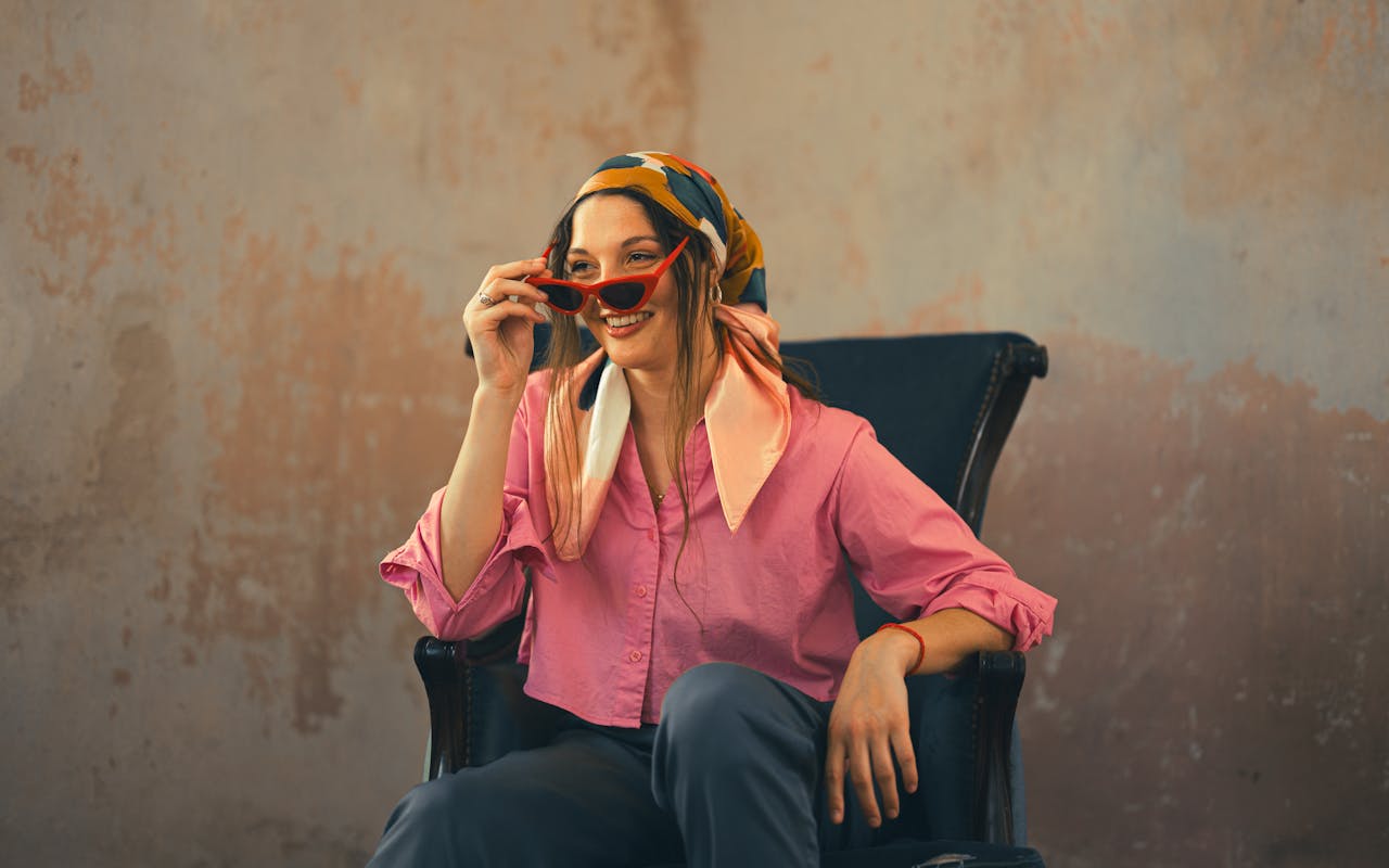 Woman in colorful attire smiles while adjusting red sunglasses in a cozy indoor setting.